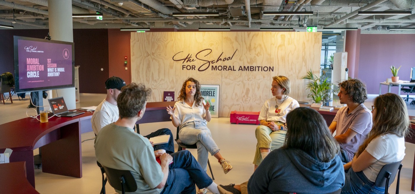A facilitator speaking to participants seated in a circle in front of a plywood wall reading The School for Moral Ambition.