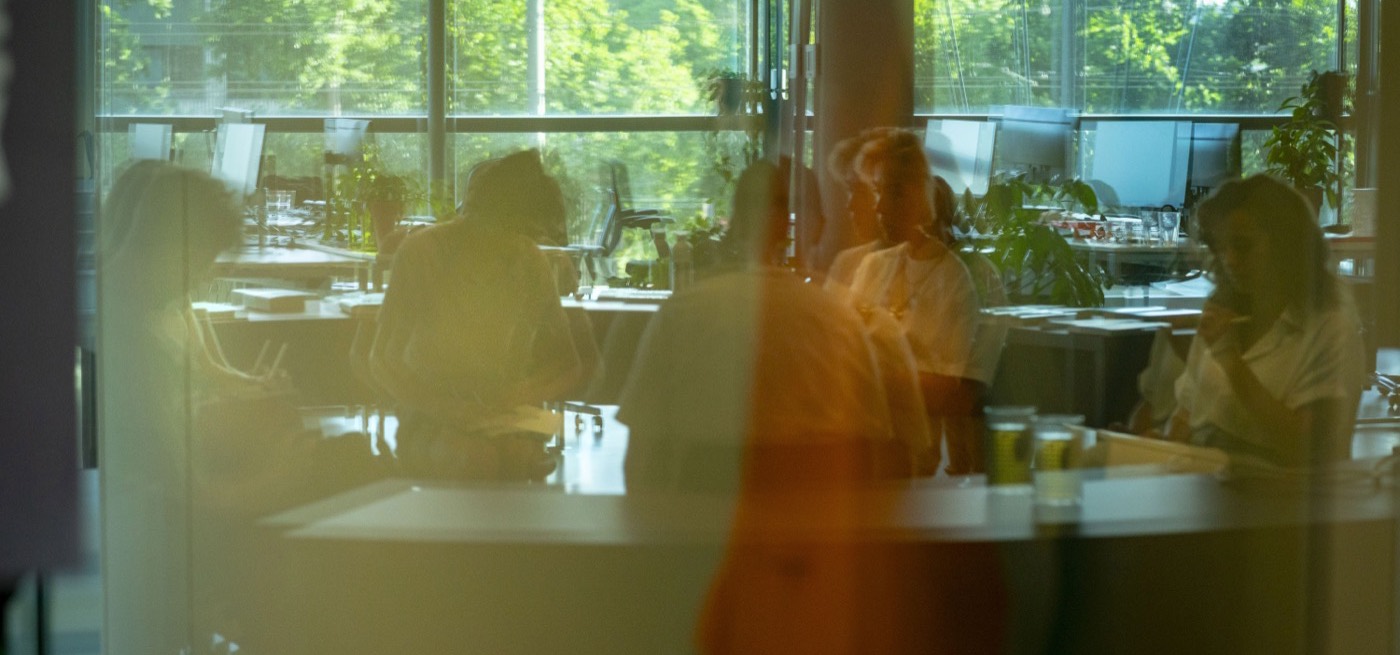 View through glass into the office, with people at desks, plants and trees outside the window.