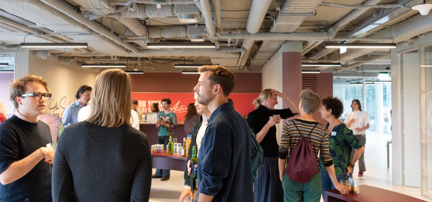 People standing in groups with drinks in the open office, in front of a red wall reading Turn your ideals into action.