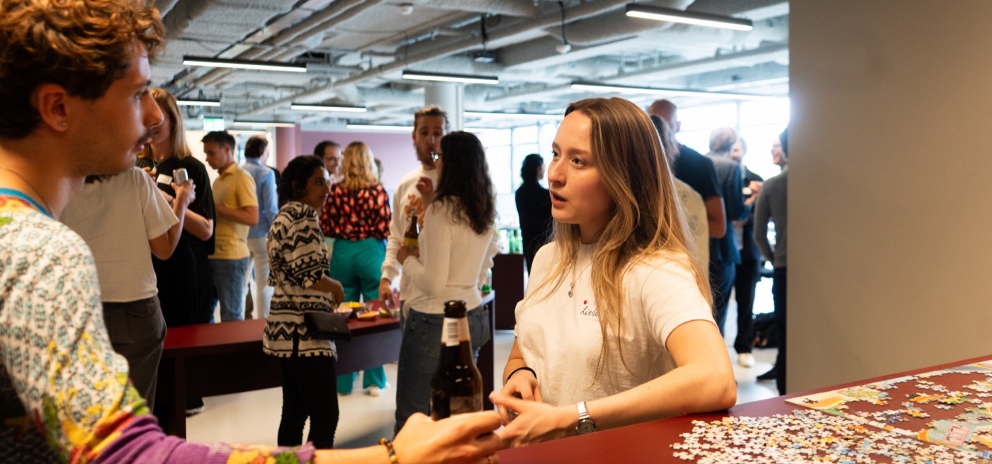 Two people in conversation across a red table with a half-finished jigsaw puzzle on it during community drinks.