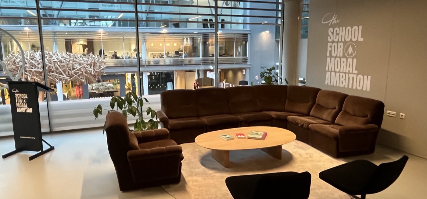Office lounge with brown sofas, a round coffee table and the School for Moral Ambition logo on the wall, looking out over the WTC atrium.